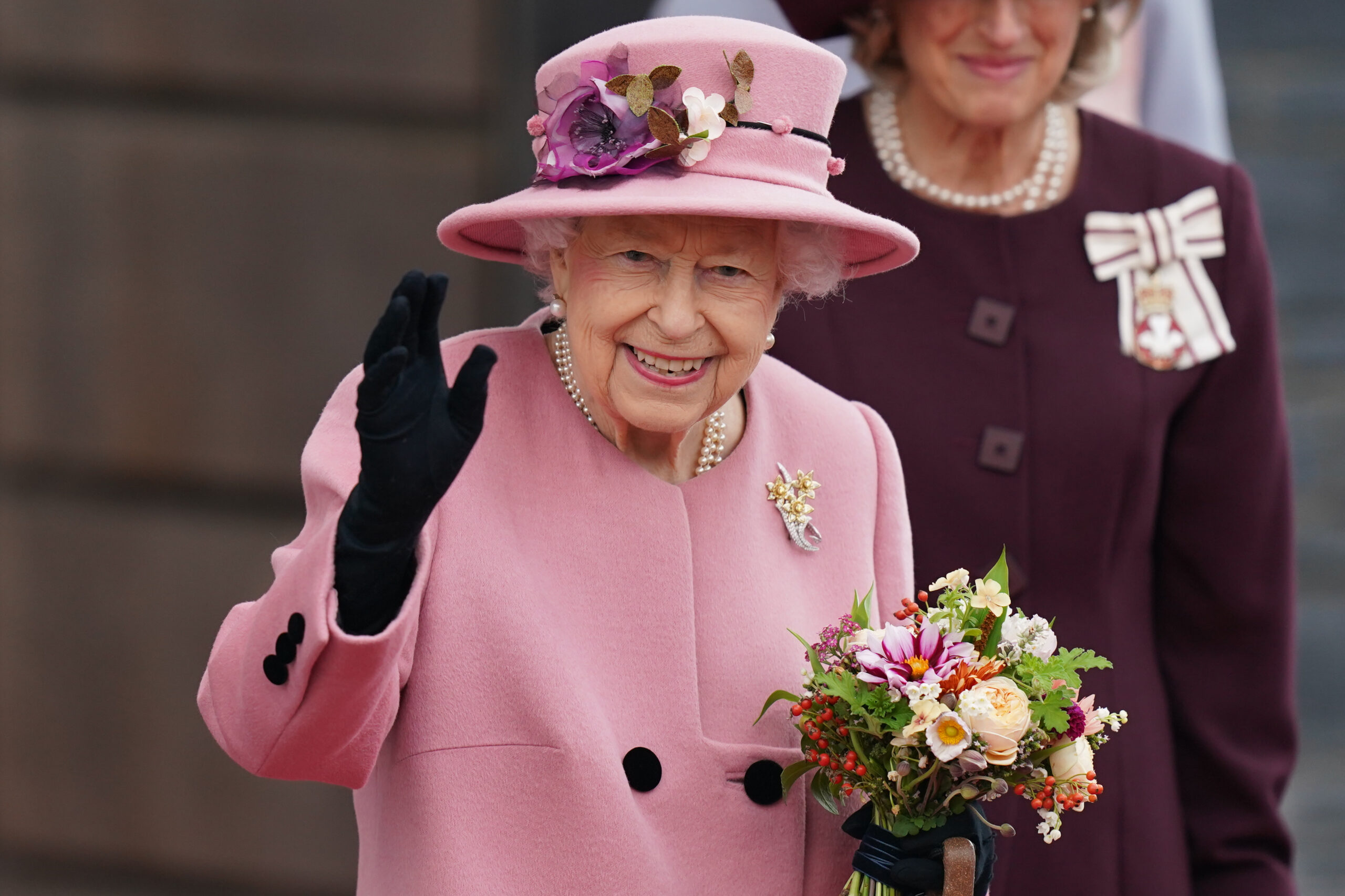 HM The Queen waving to a crowd in a pink dress, with a floral bouquet
